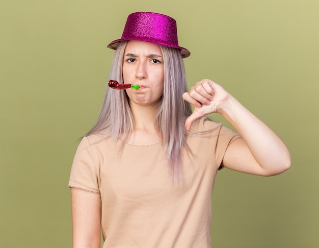 Una giovane bella ragazza scontenta che indossa il cappello da festa che soffia il fischio della festa che mostra il pollice verso il basso