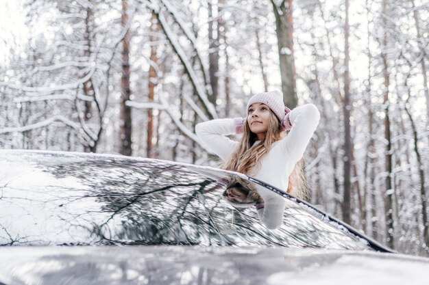 Una giovane bella ragazza vestita con un cappello leggero invernale e un maglione bianco guarda fuori dal finestrino dell'auto in una foresta innevata d'inverno