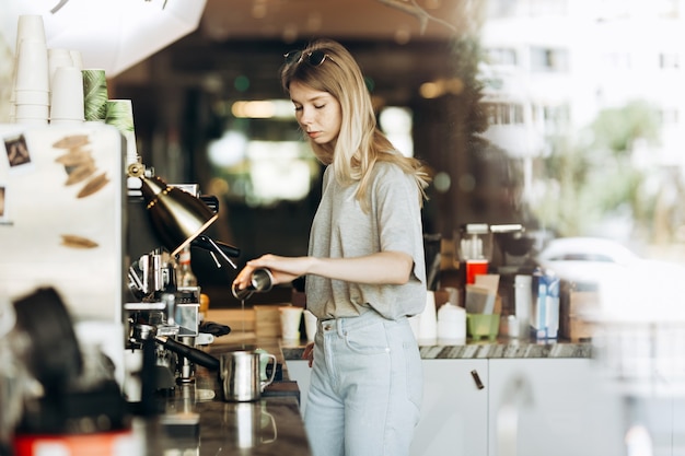 Una giovane bionda piuttosto magra con i capelli lunghi, vestita in modo casual, sta cucinando il caffè in una moderna caffetteria. Viene mostrato il processo di preparazione del caffè. .