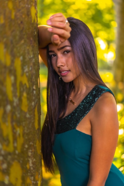 Una giovane bruna latina con lunghi capelli lisci appoggiata a un albero in un vestito verde
