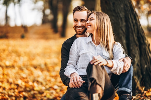 Una giovane coppia amorosa che si siede su un banco di legno nella foresta. Uomo e donna che abbracciano e che sorridono sui precedenti degli alberi di autunno