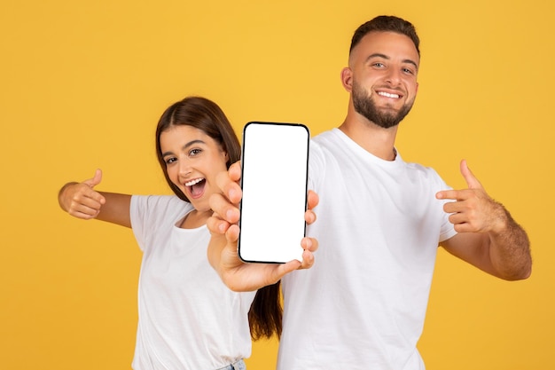 Una giovane coppia europea sorridente e sicura in magliette bianche indica con le dita un telefono con lo schermo vuoto