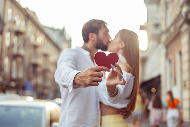 Una giovane coppia innamorata tiene un cuore rosso mostra il loro amore e si bacia in città concetto romantico