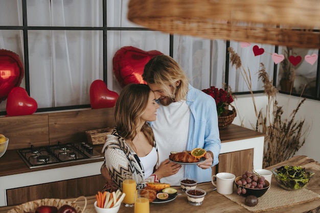 Una giovane coppia sorridente che fa colazione in cucina con palloncini a forma di cuore rosso sullo sfondo