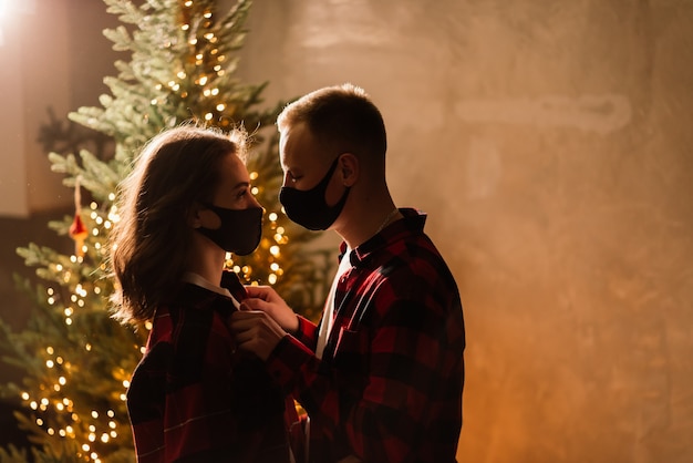 Una giovane coppia uomo e donna in maschere mediche usa e getta sotto un albero di Natale.