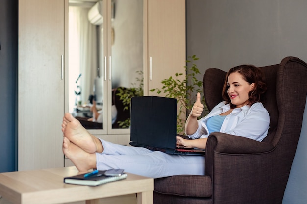 Una giovane di 30 anni in camicia bianca siede a casa su una comoda poltrona e comunica con i parenti usando un computer portatile e una videochiamata.
