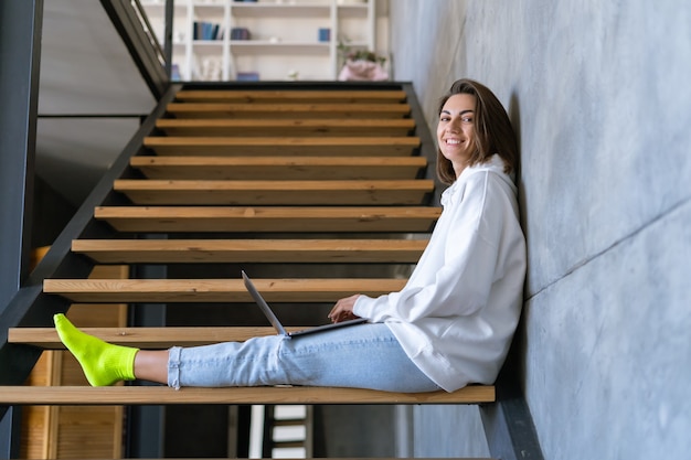 Una giovane donna a casa con una felpa con cappuccio bianca e jeans si siede sulle scale con un laptop sulle ginocchia, lavora da remoto, studia