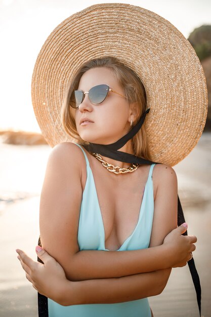 Una giovane donna abbronzata in un bellissimo costume da bagno con un cappello di paglia si alza e riposa su una spiaggia tropicale con sabbia e guarda il tramonto e il mare. Messa a fuoco selettiva. Concetto di vacanza al mare