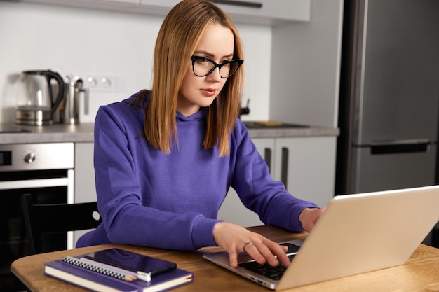 Una giovane donna adulta pensierosa con gli occhiali lavora nello studio in cucina usando l'istruzione remota del laptop