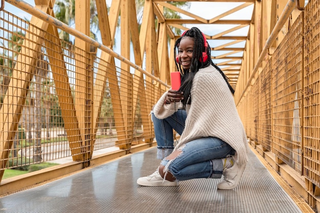 Una giovane donna afroamericana in trecce che ascolta musica con le cuffie accovacciate su un ponte giallo