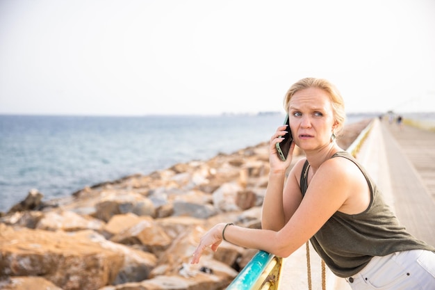 Una giovane donna alla moda comunica su uno smartphone sull'argine della città Riposa e lavora nella natura in riva al mare