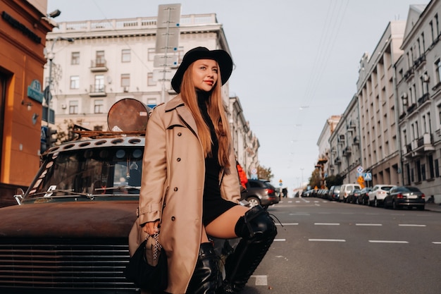 Una giovane donna alla moda con un cappotto beige e un cappello nero su una strada cittadina si siede sul cofano di un'auto al tramonto. Moda di strada femminile. Abbigliamento autunnale Stile urbano.