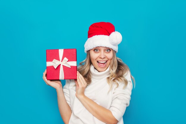 Una giovane donna allegra con un maglione bianco Cappello da Babbo Natale mostra la confezione regalo con un fiocco bianco