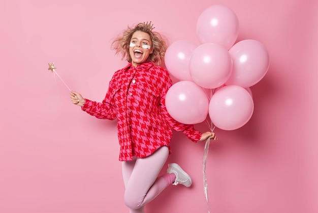 Una giovane donna allegra e ottimista balla con un mazzo di palloncini di elio viene a congratularsi con l'amico con l'anniversario esclama con gioia tiene la bacchetta magica indossa i cerotti di bellezza sotto gli occhi prima della festa