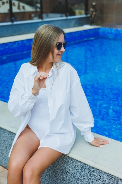 Una giovane donna allegra in costume da bagno bianco e camicia bianca è seduta a bordo piscina Una donna con occhiali da sole e costume da bagno Vacanze estive in piscina Ragazza abbronzata