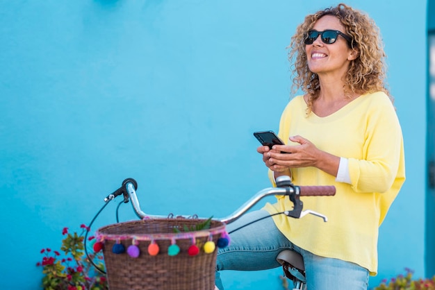 Una giovane donna allegra sorride e si diverte utilizzando la connessione del telefono cellulare Persone femminili moderne che chiacchierano all'aperto Sfondo muro blu e indumento casual giallo Bici e tempo libero Trasporto verde