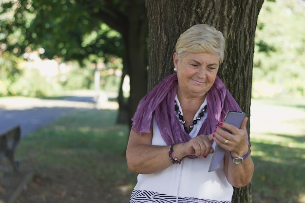 Una giovane donna anziana con uno smartphone si appoggia all'albero del parco