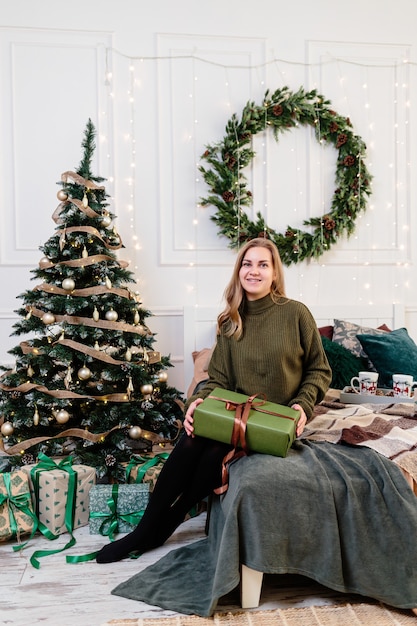 Una giovane donna apre i regali di Natale in un'atmosfera di Capodanno e gioisce. La donna è in una camera da letto luminosa e accogliente, splendidamente decorata per Natale.