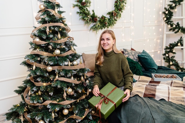 Una giovane donna apre i regali di Natale in un'atmosfera di Capodanno e gioisce. La donna è in una camera da letto luminosa e accogliente, splendidamente decorata per Natale.