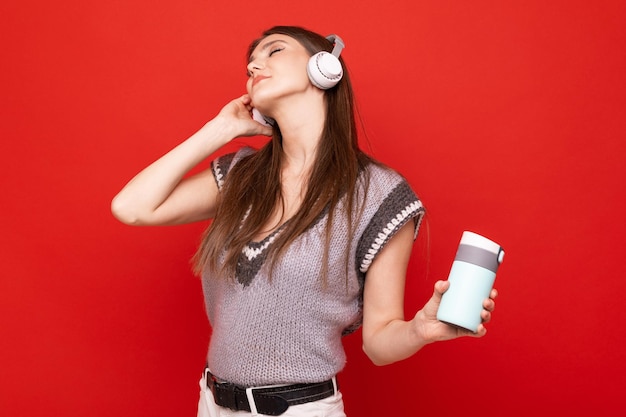 Una giovane donna ascolta musica con le cuffie e beve da una tazza termica su sfondo rosso