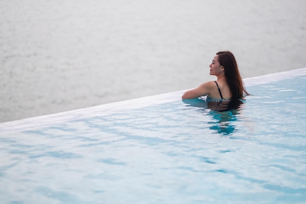 Una giovane donna asiatica che si rilassa nella piscina a sfioro guardando una bellissima vista sul mare