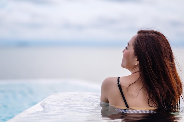 Una giovane donna asiatica che si rilassa nella piscina a sfioro guardando una bellissima vista sul mare