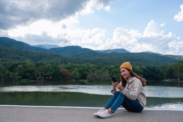 Una giovane donna asiatica che tiene e usa il telefono cellulare mentre era seduto in riva al lago e alle montagne