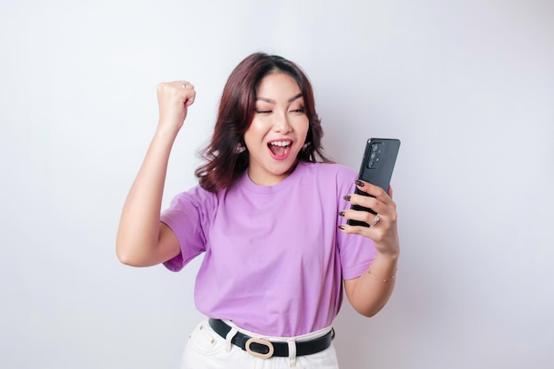 Una giovane donna asiatica con una felice espressione di successo che indossa una camicia viola lilla e tiene uno smartphone isolato da uno sfondo bianco