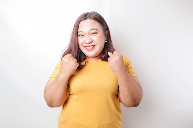 Una giovane donna asiatica di grandi dimensioni con una felice espressione di successo che indossa una camicia gialla isolata da uno sfondo bianco