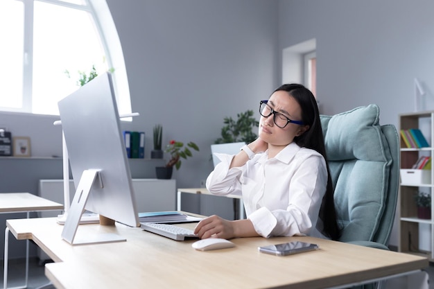 Una giovane donna asiatica impiegata in ufficio con gli occhiali e una camicia bianca si siede a una scrivania a un computer stanco