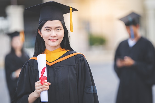 Una giovane donna asiatica laureata in abito da laurea e sparviere in possesso di un certificato di laurea si trova di fronte all'edificio universitario dopo aver partecipato all'inizio del college