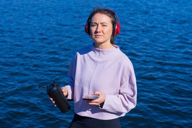 Una giovane donna atletica ascolta musica e beve acqua durante le pause dall'allenamento all'aperto