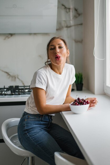 Una giovane donna attraente mangia ciliegie fresche bacche sane seduta a casa in cucina e scherzando