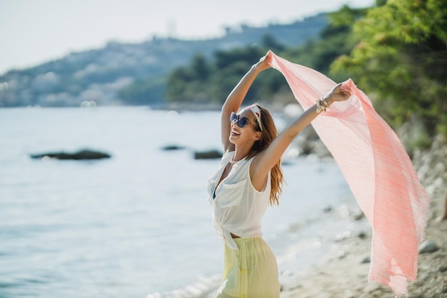 Una giovane donna attraente si diverte e si gode le vacanze estive. Sta posando con una sciarpa trasparente tra le mani sulla spiaggia.