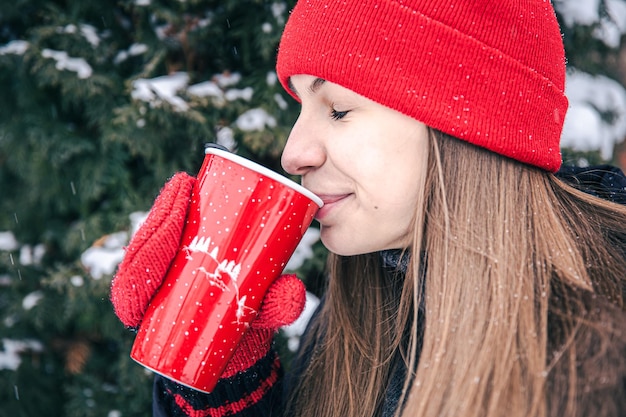 Una giovane donna beve una bevanda calda da una tazza termica rossa in inverno