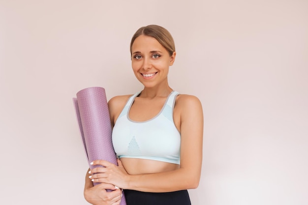 Una giovane donna bionda abbronzata adatta sorridente caucasica che tiene la stuoia di yoga nelle sue mani