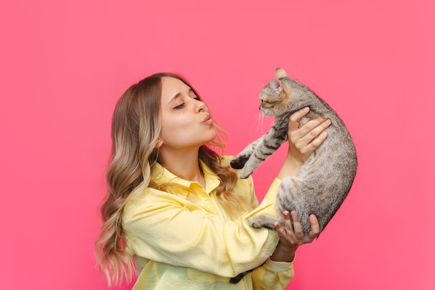 Una giovane donna bionda carina caucasica con una camicia gialla tiene in mano un gatto ammirandolo isolato su un muro rosa di colore brillante