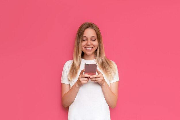 Una giovane donna bionda carina sorridente caucasica tiene un telefono cellulare guardando lo schermo isolato sul muro rosa di colore brillante La ragazza in una maglietta bianca usa il suo smartphone