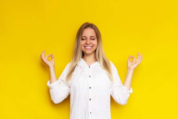 Una giovane donna bionda caucasica consapevole in una camicia bianca tiene le mani nel gesto mudra con gli occhi chiusi isolata sul muro di colore giallo La ragazza pacifica medita tenendo le dita nel segno dello yoga