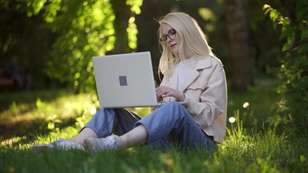 Una giovane donna bionda con un computer portatile si siede su un prato verde nel parco. Lavoro per piacere, lavoro a distanza, creatività. 4K UHD