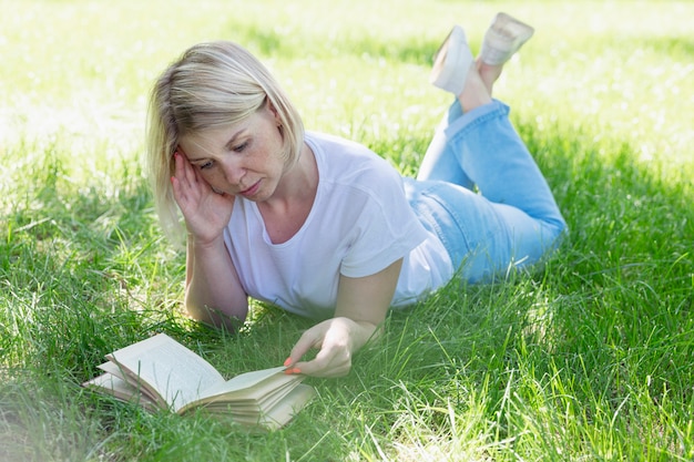 Una giovane donna bionda è sdraiata sull'erba in un parco in una soleggiata giornata estiva e sta leggendo un libro. Ricreazione e apprendimento a distanza in natura.