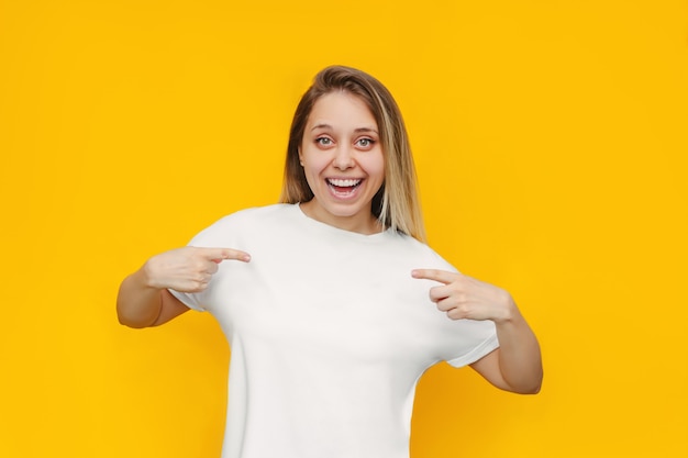 Una giovane donna bionda sorridente allegra bella caucasica che punta alla maglietta casual bianca con le dita che mostra lo spazio vuoto della copia su una parete gialla di colore brillante