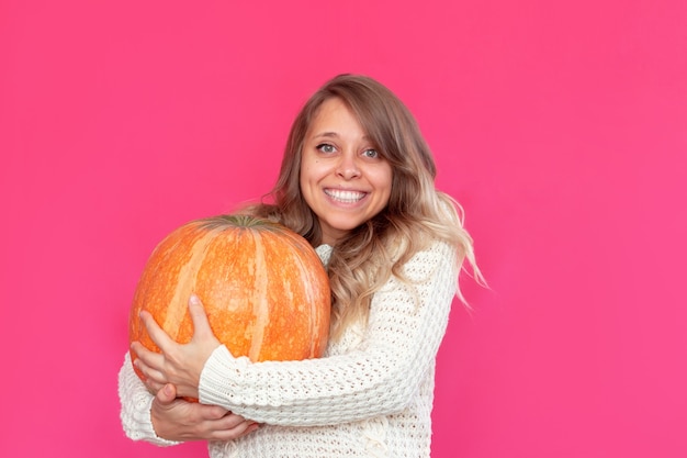 Una giovane donna bionda sorridente con un maglione caldo di lana bianca tiene una grande zucca Halloween concept