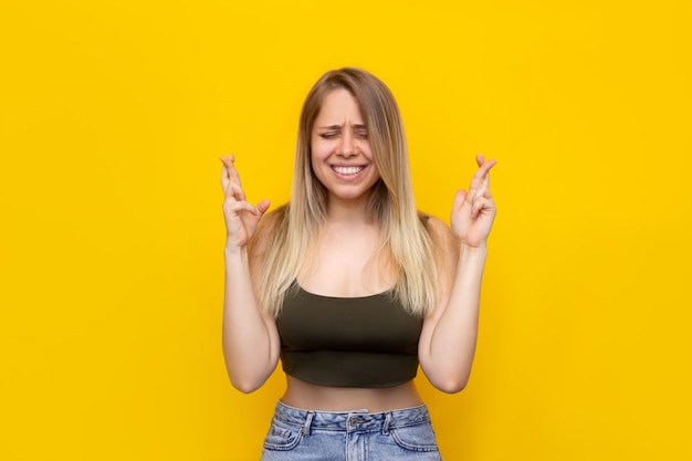 Una giovane donna bionda sorridente piuttosto caucasica con gli occhi chiusi incrocia le dita per buona fortuna in attesa dei risultati della lotteria o degli esami isolati su un muro giallo di colore brillante