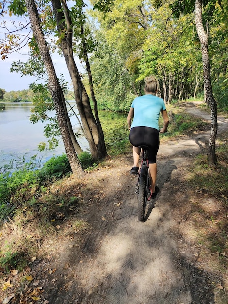 Una giovane donna bionda va in bicicletta nella foresta vicino al fiume Vista posteriore Turismo e sport