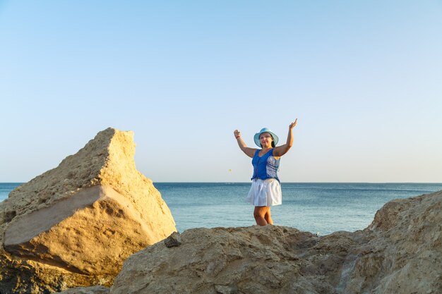 Una giovane donna bruna con una gonna bianca e un cappello da sole in riva al mare si erge sullo sfondo del mare e del cielo.