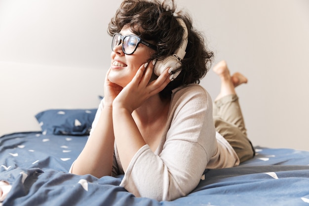 una giovane donna carina allegra si trova al chiuso sul letto ascoltando musica con le cuffie.