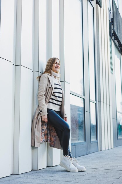 Una giovane donna carina in un impermeabile beige e stivali di pelle autunnali cammina per strada Immagine calda autunno Moda femminile