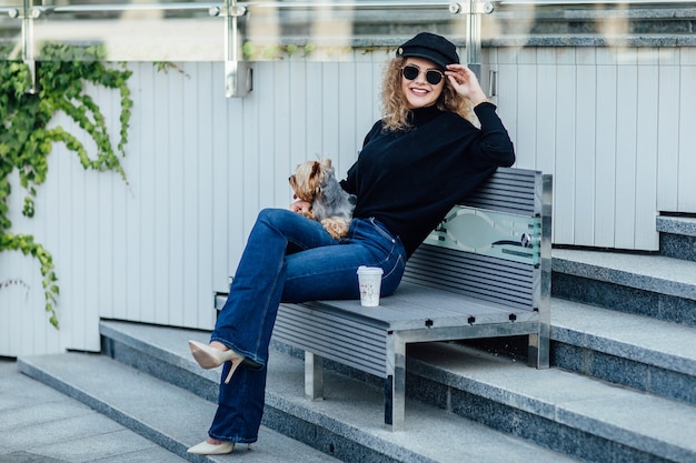 Una giovane donna caucasica che cammina lungo la strada europea con un piccolo cane di razza Chihuahua di due colori al guinzaglio Nuvoloso caldo autunno primavera tempo Ragazza vestita con camicia nera e scarpe nude