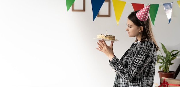 Una giovane donna caucasica si erge vestita con un abito femminile tiene una torta nelle sue mani e la guarda Concetto di compleanno in ufficio Baner